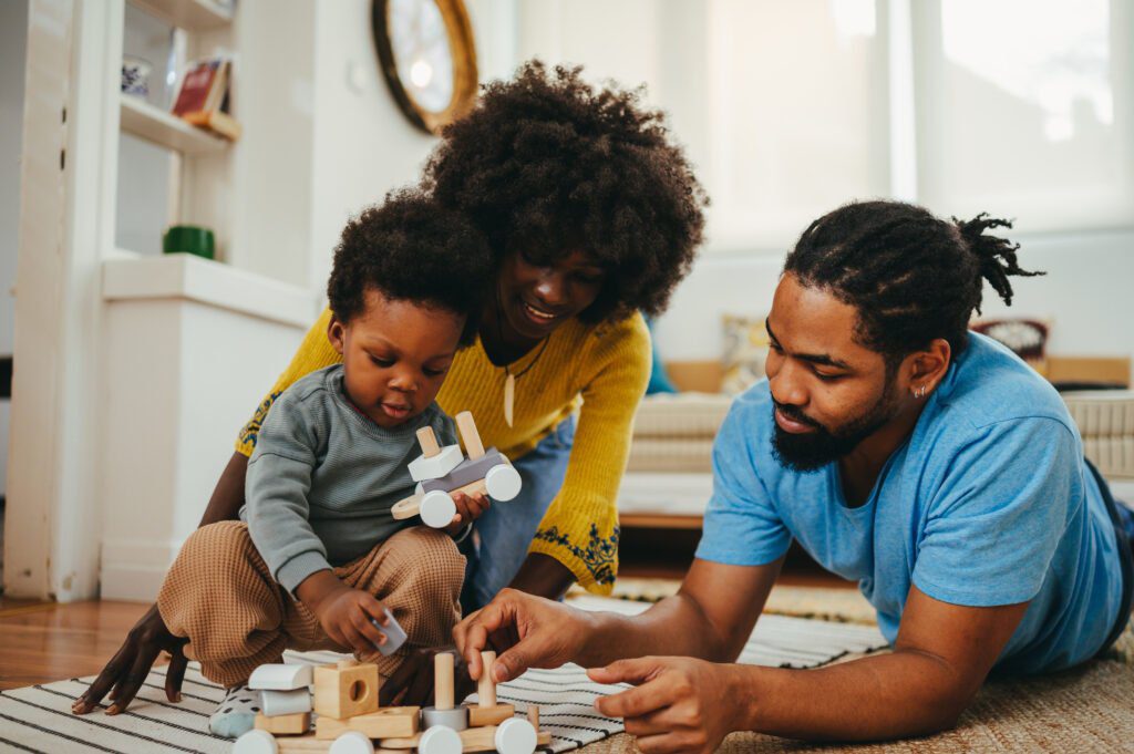 Happy african american parents are teaching their beloved little boy through the educational Montessori toy while sitting and lying on the floor in the living room. A happy family having fun together.